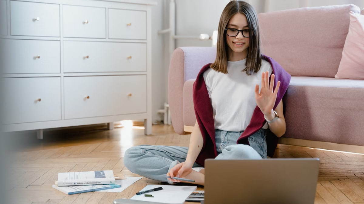 Woman studying online comfortably from home, waving during a video call.